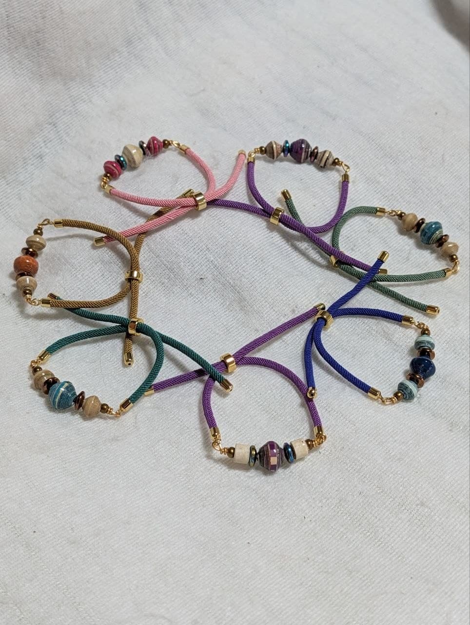 Assorted handmade Sela Bracelets with recycled paper beads on pink, purple, teal and blue braided cords.